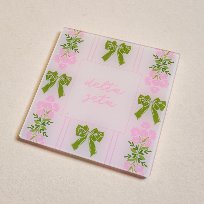 The Darling Effect’s Delta Zeta Acrylic Coaster features a light beige background, pink stripes, green bows, floral designs, and “Delta Zeta” in pink cursive—perfect for chapter pride or as a Big/Little gift.