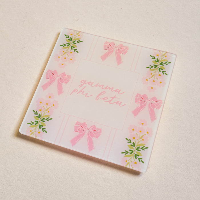 The Darling Effect's Gamma Phi Beta Acrylic Coaster features pink script, pink bows, and yellow flowers with green leaves on a light background—an adorable sorority gift.
