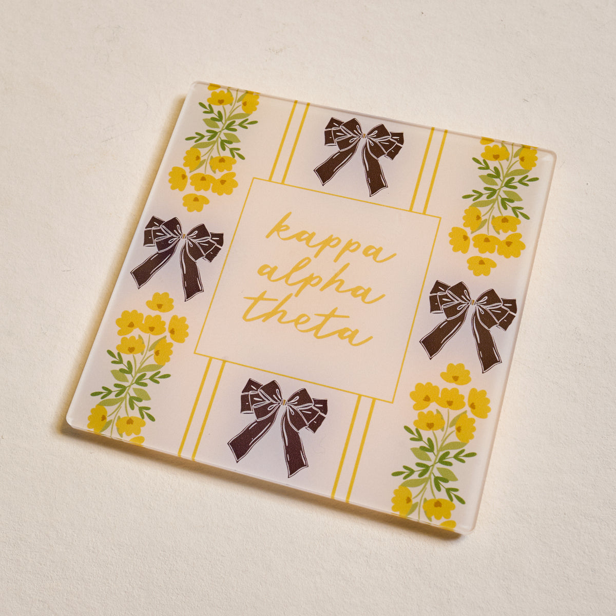 The Darling Effect’s Kappa Alpha Theta Acrylic Coaster features gold script, yellow flowers, and black bows on a cream background—perfect for chapter pride or as a special Big/Little gift.