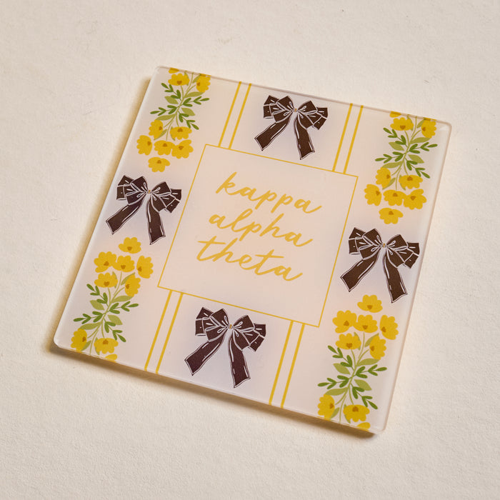The Darling Effect’s Kappa Alpha Theta Acrylic Coaster features gold script, yellow flowers, and black bows on a cream background—perfect for chapter pride or as a special Big/Little gift.