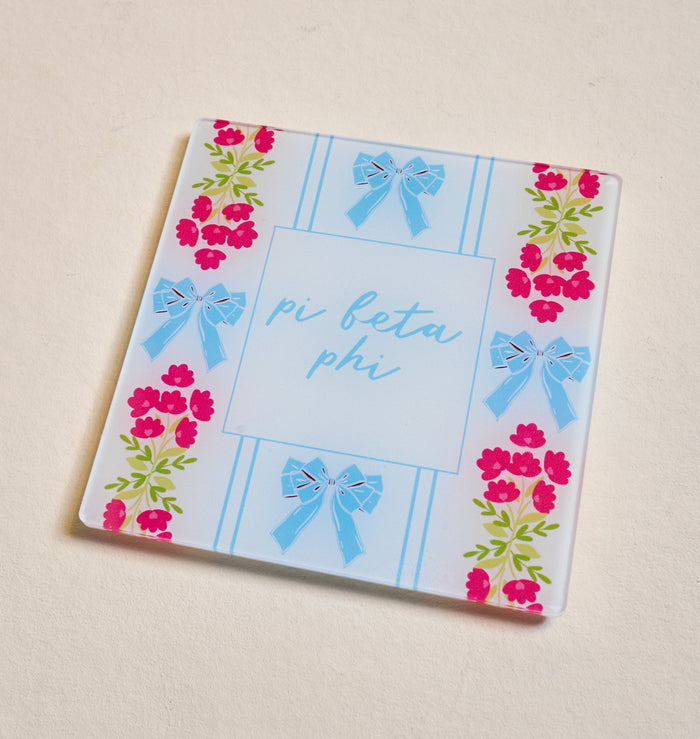 The Darling Effect’s Pi Beta Phi Acrylic Coaster features blue cursive lettering, blue bows, and pink flowers—an ideal sorority coaster or Big/Little gift—shown here on a light beige surface.