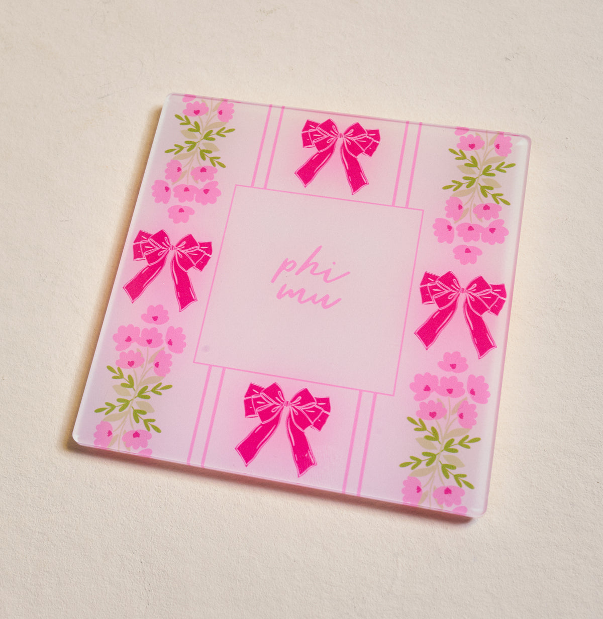 The Darling Effect's Phi Mu Acrylic Coaster is a square pink coaster featuring "phi mu" in the center with bows and floral edges—ideal for chapter pride or as a cute Big/Little gift, beautifully displayed on a light surface.