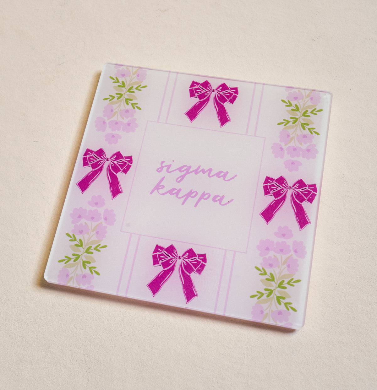 The Darling Effect’s Sigma Kappa Acrylic Coaster features pink bows and purple flowers on a light background with “Sigma Kappa” in cursive—an ideal sorority gift or stylish addition to your coaster collection.