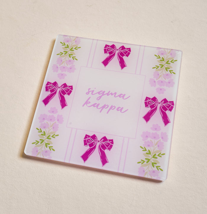 The Darling Effect’s Sigma Kappa Acrylic Coaster features pink bows and purple flowers on a light background with “Sigma Kappa” in cursive—an ideal sorority gift or stylish addition to your coaster collection.