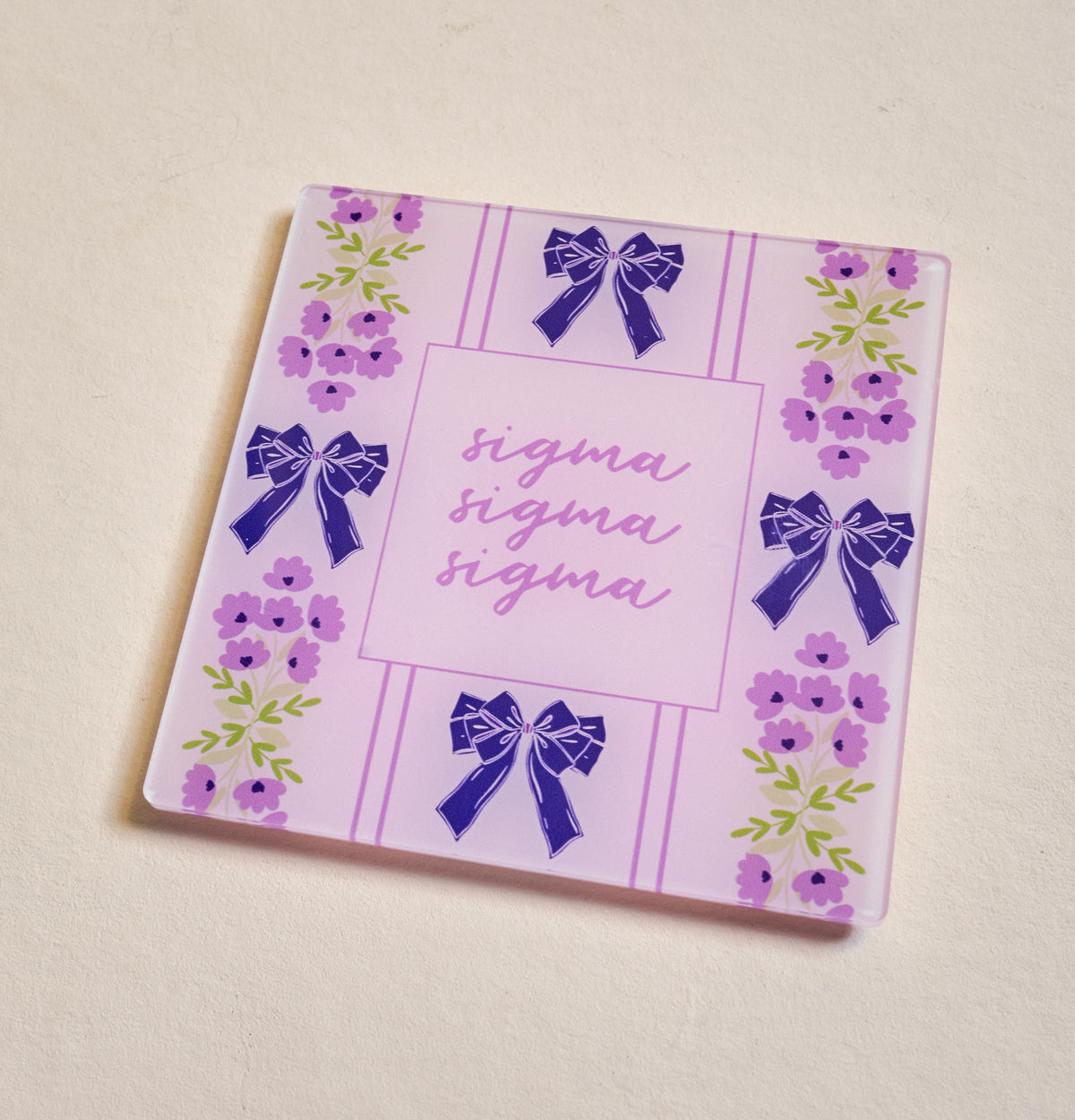 The Darling Effect's Sigma Sigma Sigma Acrylic Coaster features a pink and purple floral design with blue bows and "sigma" scripted three times—a perfect sorority gift for any occasion.