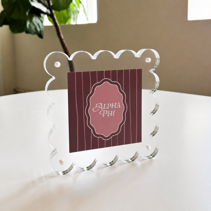 The Darling Effect’s Alpha Phi Square Scalloped Acrylic Frame, featuring a clear scalloped-edge design, displays a pink and maroon Alpha Phi card. It sits magnetized on a white table near a green plant with windows in the background.