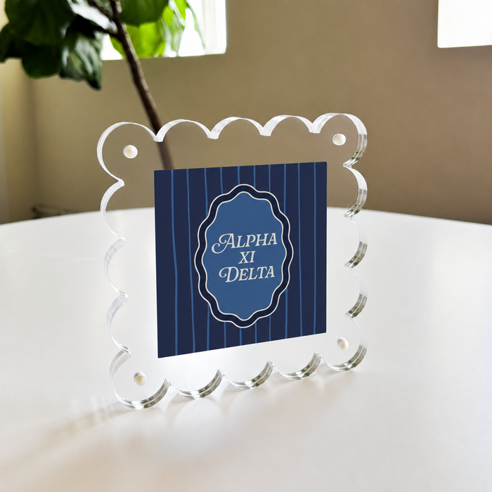 The Darling Effect Alpha Xi Delta Square Scalloped Acrylic Frame features a blue card with "Alpha Xi Delta" in white script, displayed on a white table with a plant and window in the background.