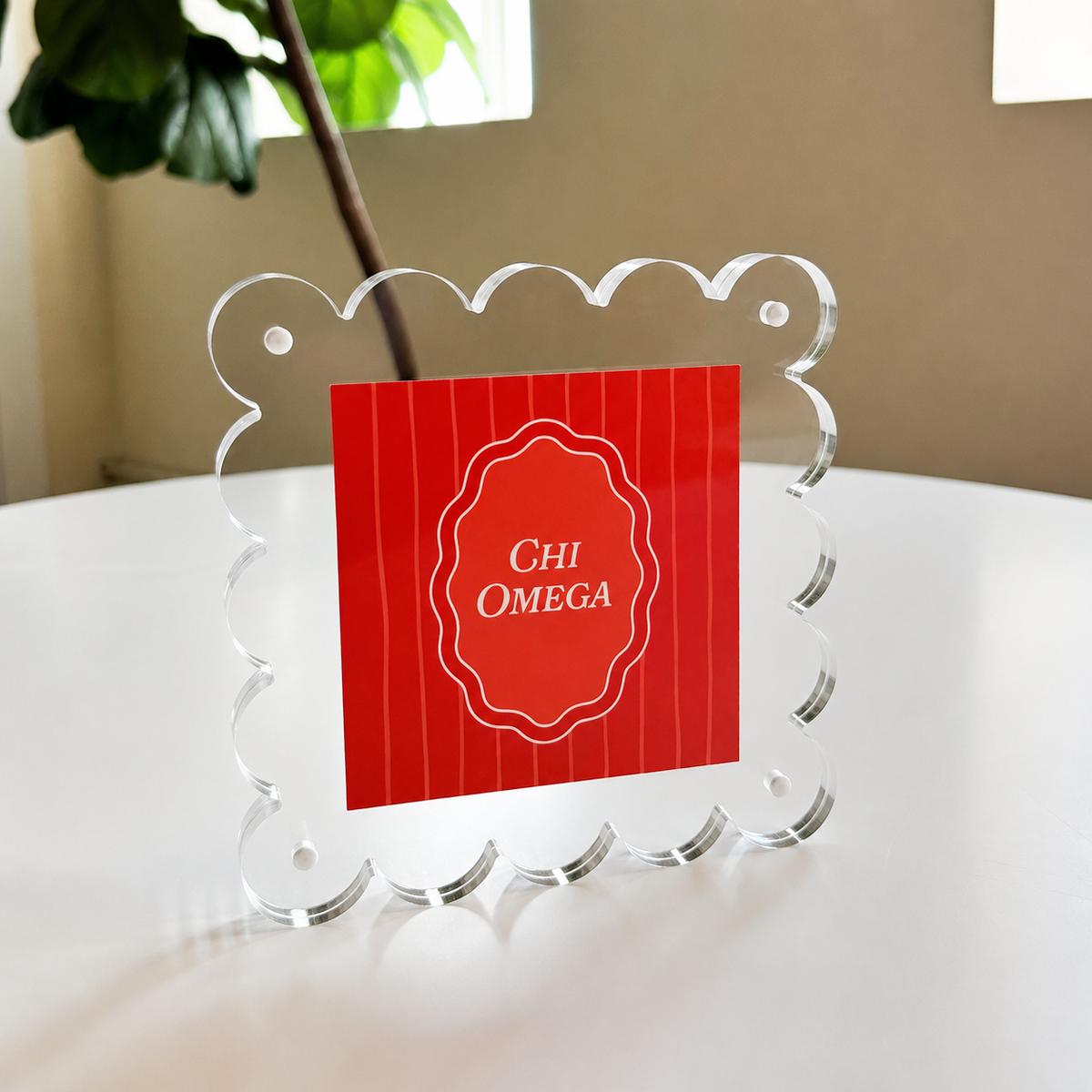 The Darling Effect's Chi Omega Square Scalloped Acrylic Frame showcases a red card with "Chi Omega" in white text, displayed on a white round table with a green plant and window in the background.