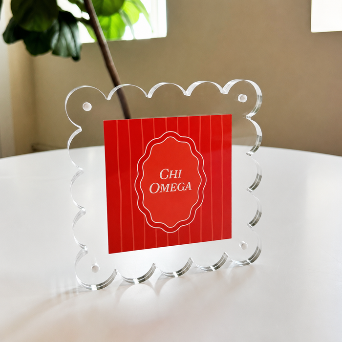 The Darling Effect's Chi Omega Square Scalloped Acrylic Frame showcases a red card with "Chi Omega" in white text, displayed on a white round table with a green plant and window in the background.