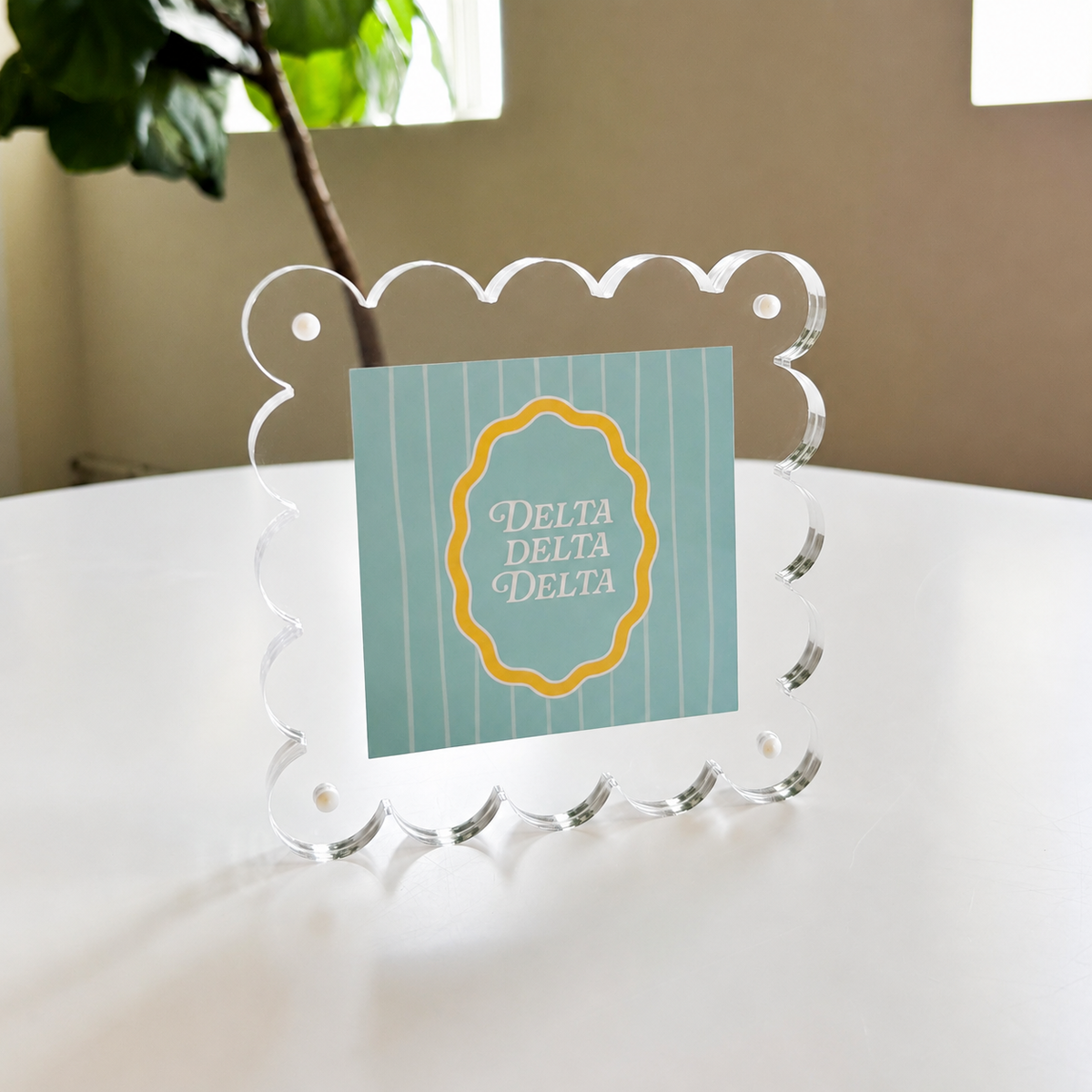 The Darling Effect’s Delta Delta Delta Square Scalloped Acrylic Frame features a modern, scalloped-edge design and displays a blue and white striped card with a yellow-edged emblem, adding charm to any space.