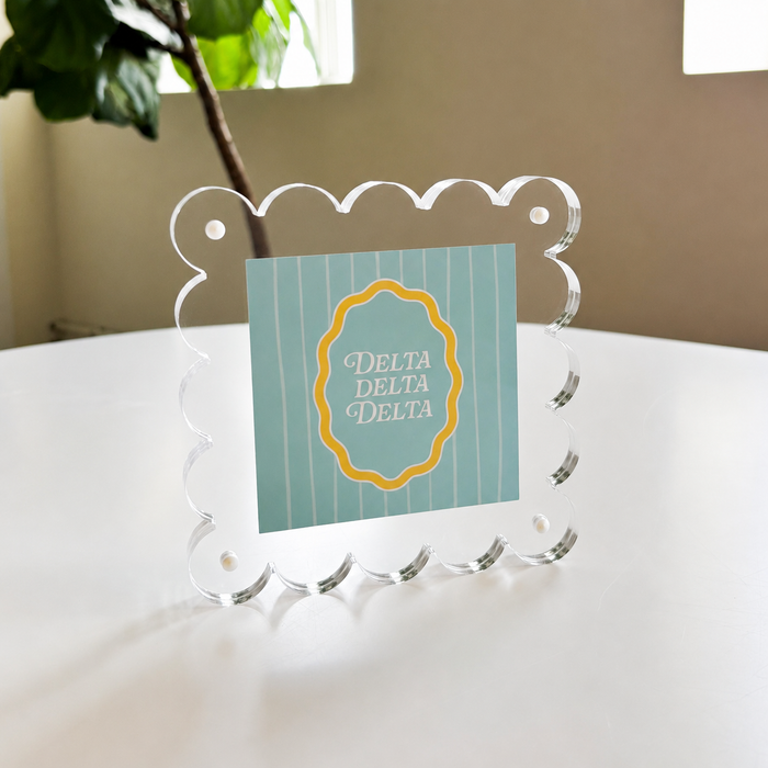 The Darling Effect’s Delta Delta Delta Square Scalloped Acrylic Frame features a modern, scalloped-edge design and displays a blue and white striped card with a yellow-edged emblem, adding charm to any space.