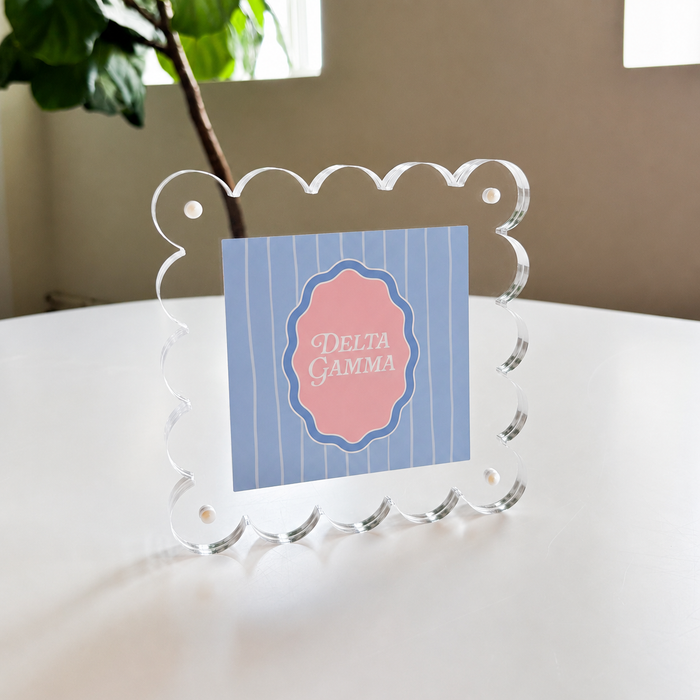 The Darling Effect's Delta Gamma Square Scalloped Acrylic Frame holds a blue and white striped card with a pink "Delta Gamma" center, displayed on a white table beside a green plant.