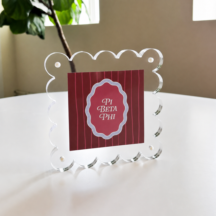 The Darling Effect's Pi Beta Phi Square Scalloped Acrylic Frame displays a red PI BETA PHI card on a white table, with leafy plants and windows behind—perfect for showcasing memories in modern style.