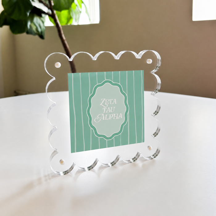 The Darling Effect’s Zeta Tau Alpha Square Scalloped Acrylic Frame displays a mint green card with white script, shown on a white table beside a leafy plant and window for modern decorative flair.