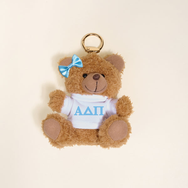 The Darling Effect's Alpha Delta Pi Teddy Bear Bag Charm is a small brown bear keychain with a blue bow and white shirt featuring blue ΑΔΠ Greek letters—perfect as a Bid Day gift or cute accessory.