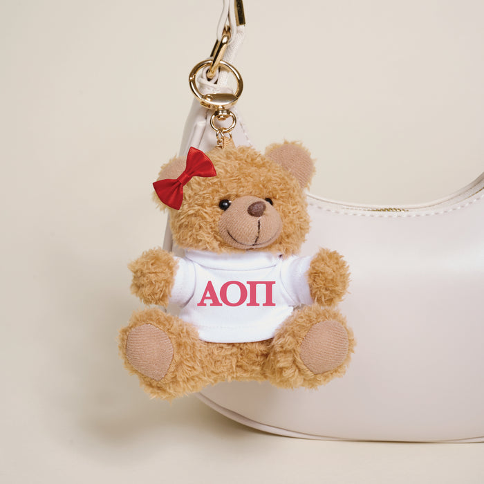 The Darling Effect Alpha Omicron Pi Teddy Bear Bag Charm features a small teddy bear with a red bow and white shirt with red ΑΟΠ letters, hanging as a cute keychain accessory on any bag.