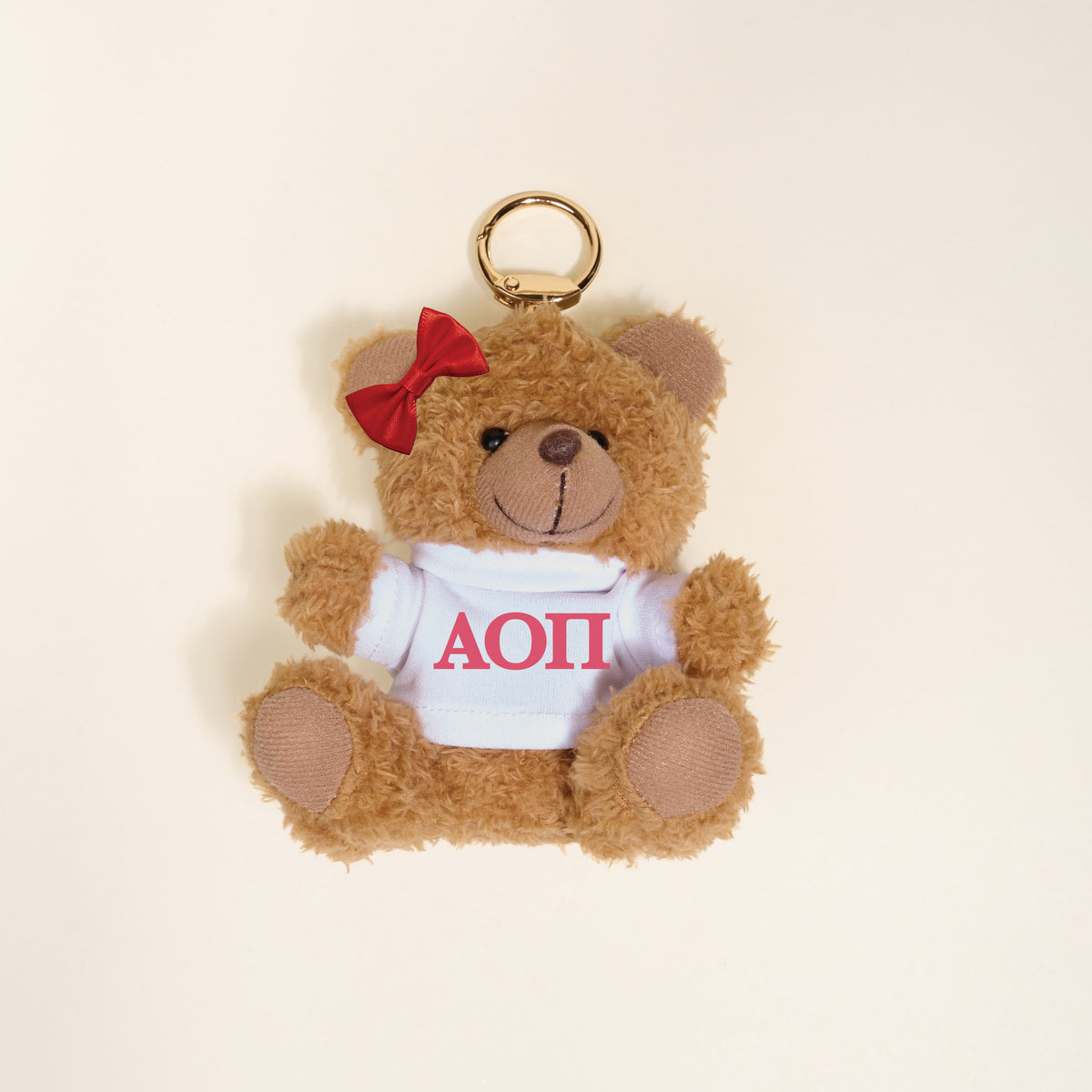 The Darling Effect's Alpha Omicron Pi Teddy Bear Bag Charm features a small brown bear keychain with a red bow and a white shirt with red Greek letters ΑΟΠ, shown on a simple light background.