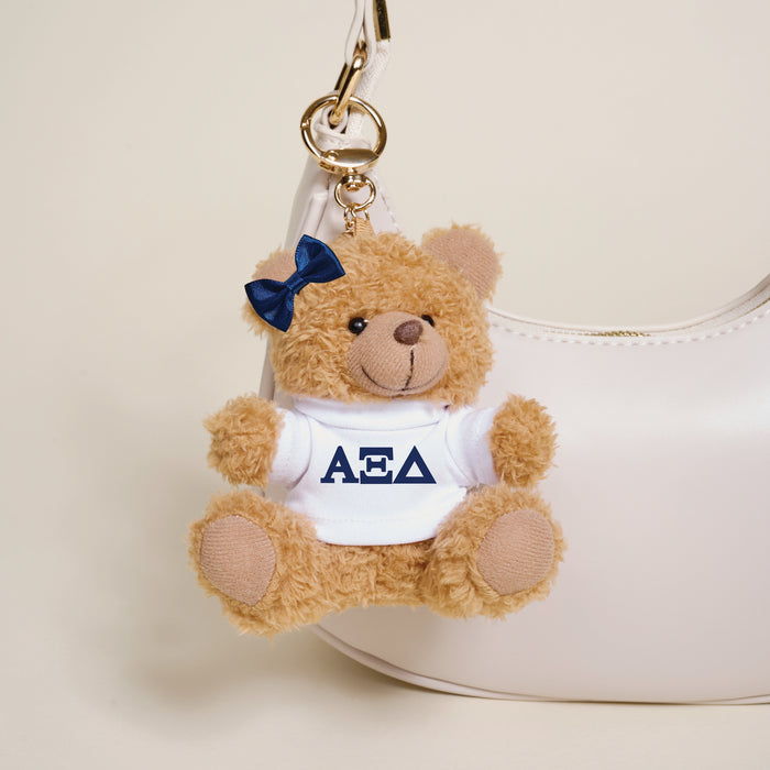 The Darling Effect’s Alpha Xi Delta Teddy Bear Bag Charm is a small brown teddy bear keychain with a navy bow and white shirt featuring Greek letters, perfect for accessorizing your white handbag.