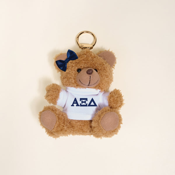 The Darling Effect’s Alpha Xi Delta Teddy Bear Bag Charm is a small brown keychain with a blue bow on its left ear and a white shirt featuring Greek letters—perfect as a sorority gift or bag accessory against a light background.