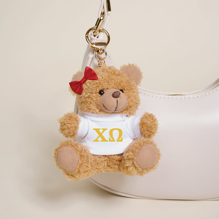 The Darling Effect's Chi Omega Teddy Bear Bag Charm—a small brown bear with a red bow and white shirt with yellow ΧΩ letters—hangs from a white handbag, making it an ideal sorority gift or cute accessory.