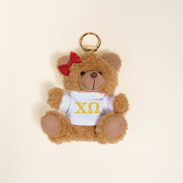 The Darling Effect Chi Omega Teddy Bear Bag Charm features a cute mini bear with a red bow and white shirt with yellow ΧΩ Greek letters—perfect as a sorority gift or bag charm. Displayed on a simple light background.