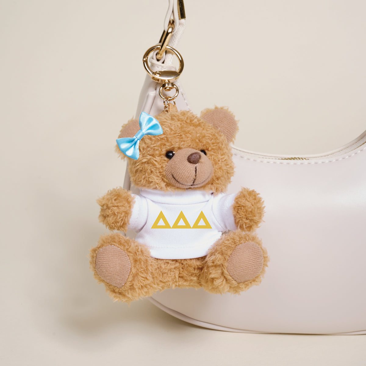 A small brown Delta Delta Delta Teddy Bear Bag Charm by The Darling Effect, with a blue bow and white shirt featuring three yellow triangles, hangs from a white handbag.