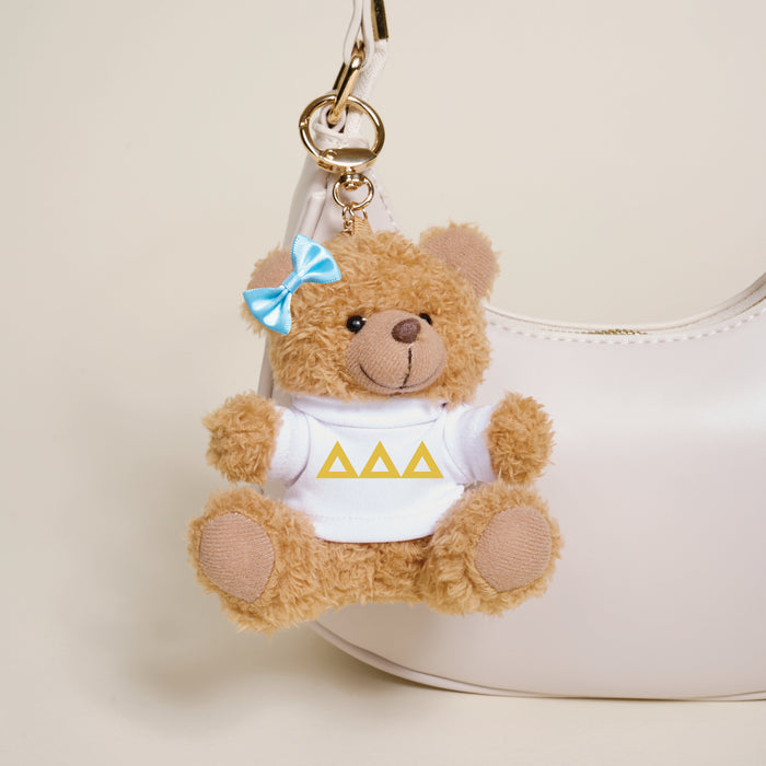 A small brown Delta Delta Delta Teddy Bear Bag Charm by The Darling Effect, with a blue bow and white shirt featuring three yellow triangles, hangs from a white handbag.