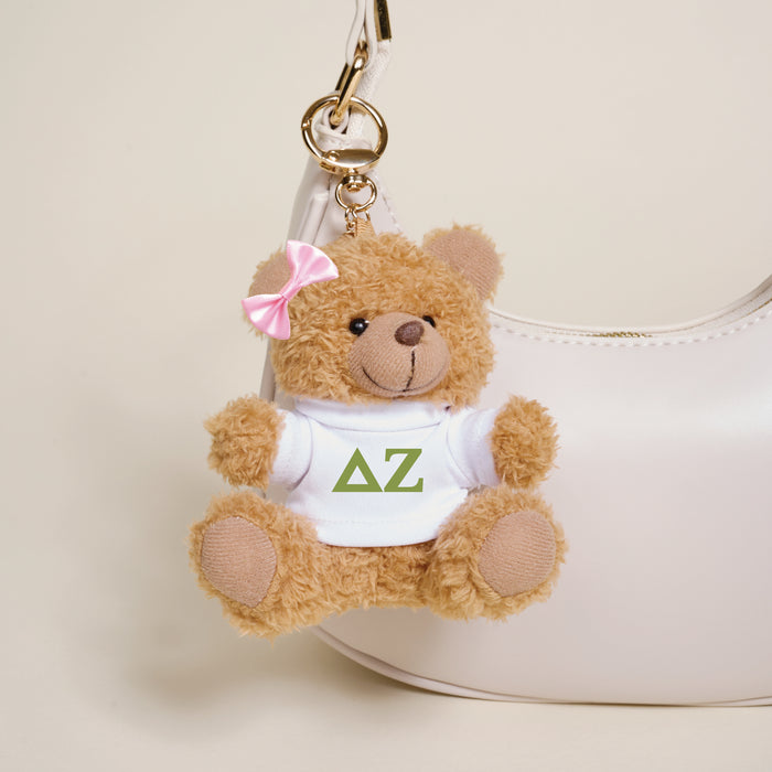 The Darling Effect's Delta Zeta Teddy Bear Bag Charm—a small brown bear with a pink bow and white shirt with green letters—is attached to a white handbag, making it a perfect accessory or Bid Day gift.