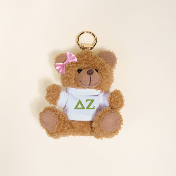 The Darling Effect’s Delta Zeta Teddy Bear Bag Charm features a small brown bear with a pink bow, wearing a white shirt with green ΔΖ letters—perfect as a Bid Day gift or cute accessory for your bag.