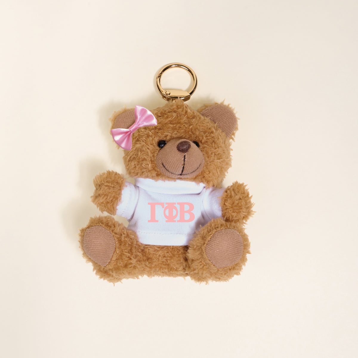 A small brown Gamma Phi Beta Teddy Bear Bag Charm by The Darling Effect, with a pink bow and white shirt with pink Greek letters, shown on a light beige background—ideal as a cute sorority accessory or bag charm.