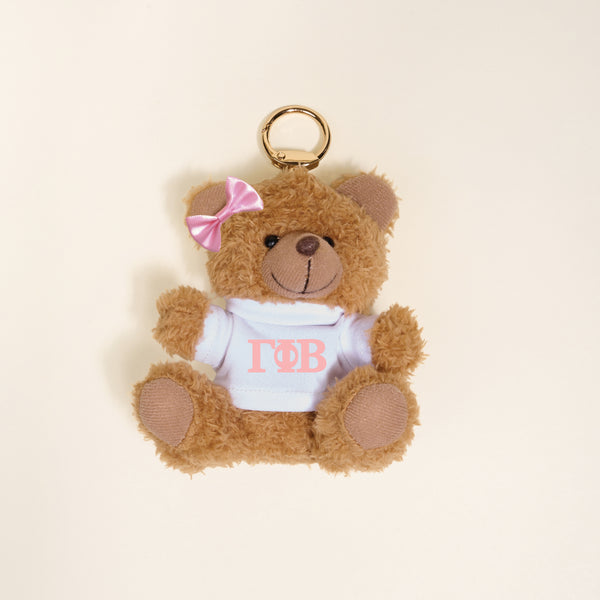 A small brown Gamma Phi Beta Teddy Bear Bag Charm by The Darling Effect, with a pink bow and white shirt with pink Greek letters, shown on a light beige background—ideal as a cute sorority accessory or bag charm.