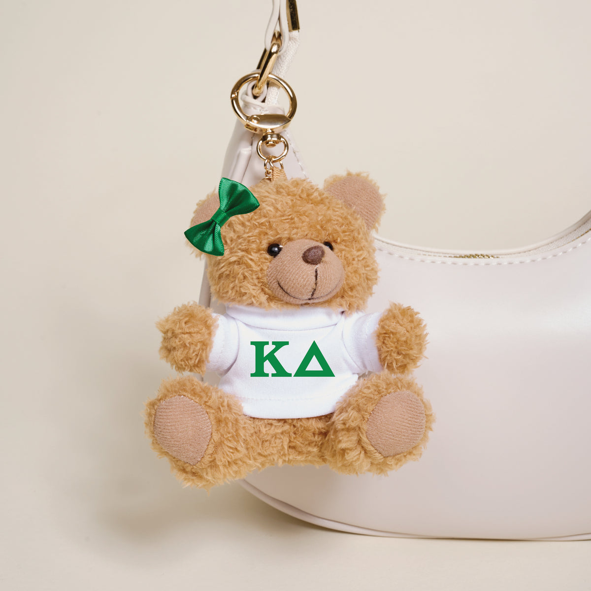 The Darling Effect Kappa Delta Teddy Bear Bag Charm—a small brown bear with a green bow and a white shirt with green KΔ letters—hangs from a white handbag, making the perfect sorority gift or accessory.