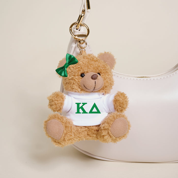 The Darling Effect Kappa Delta Teddy Bear Bag Charm—a small brown bear with a green bow and a white shirt with green KΔ letters—hangs from a white handbag, making the perfect sorority gift or accessory.