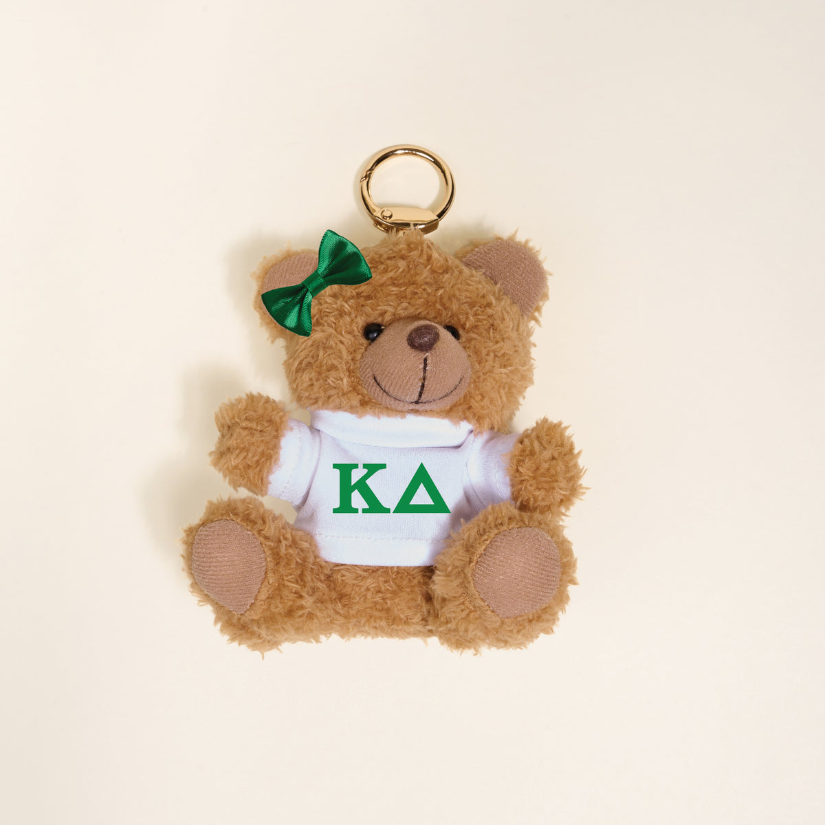 The Darling Effect’s Kappa Delta Teddy Bear Bag Charm features a small brown bear with a green bow on its left ear, wearing a white shirt with green KΔ Greek letters—perfect as a sorority gift or keychain accessory. Photographed on a light background.
