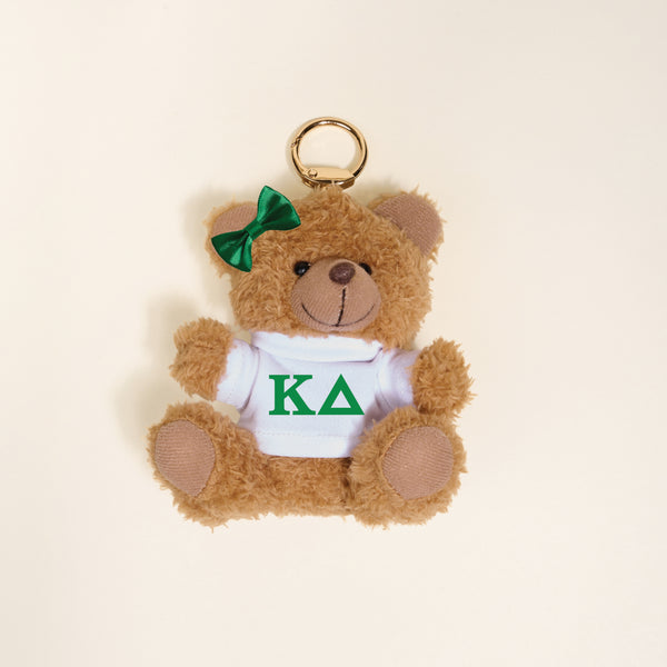 The Darling Effect’s Kappa Delta Teddy Bear Bag Charm features a small brown bear with a green bow on its left ear, wearing a white shirt with green KΔ Greek letters—perfect as a sorority gift or keychain accessory. Photographed on a light background.