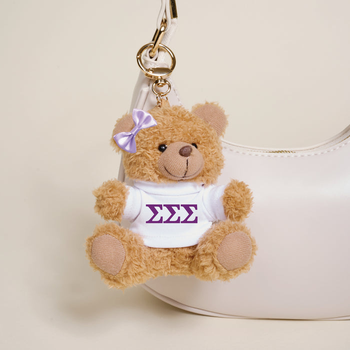 The Darling Effect’s Sigma Sigma Sigma Teddy Bear Bag Charm, a small brown bear with a purple bow and white ΣΣΣ shirt, hangs from a white handbag—an ideal sorority gift or accessory.