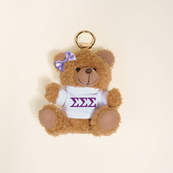 The Darling Effect’s Sigma Sigma Sigma Teddy Bear Bag Charm features a small brown teddy bear with a white shirt displaying purple ΣΣΣ Greek letters and a matching purple bow, perfect as a sorority gift or bag accessory.
