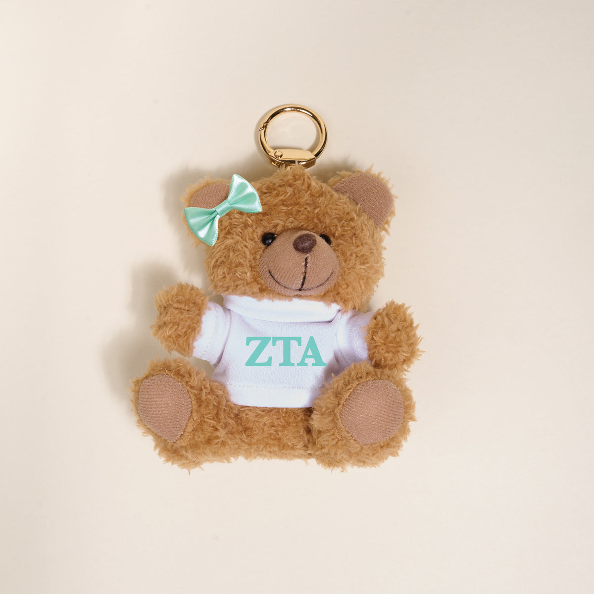 The Darling Effect Zeta Tau Alpha Teddy Bear Bag Charm is a small brown plush keychain with a teal bow and white shirt with teal ZTA letters—perfect for Bid Day baskets or as a cute bag accessory. Set against a light beige background.