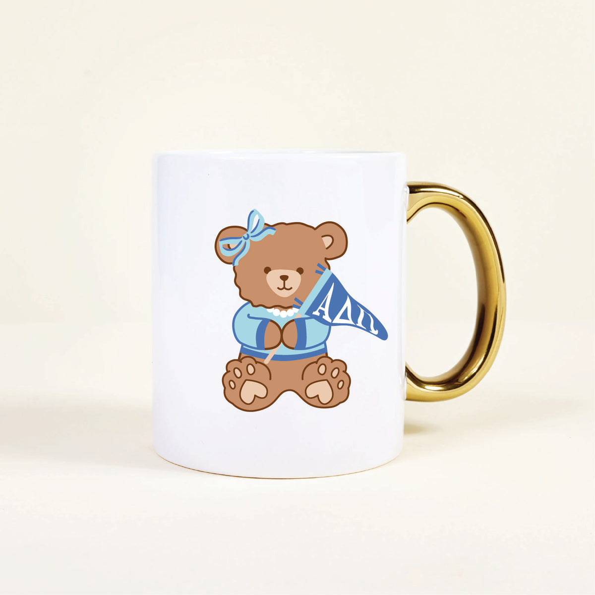 The Darling Effect’s Alpha Delta Pi 11 oz Cozy Cute Ceramic Mug features a brown teddy bear in a blue bow and sweater holding an ΑΔΠ pennant, plus a shiny gold handle for extra charm.