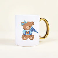 The Darling Effect’s Alpha Delta Pi 11 oz Cozy Cute Ceramic Mug features a brown teddy bear in a blue bow and sweater holding an ΑΔΠ pennant, plus a shiny gold handle for extra charm.