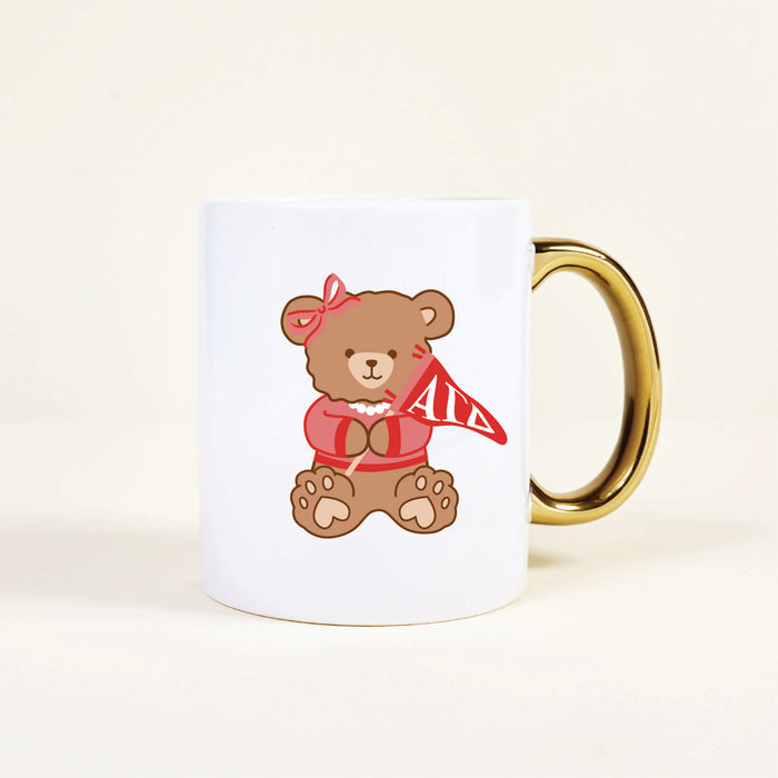 The Darling Effect's Alpha Gamma Delta 11 oz Cozy Cute Ceramic Mug features a ceramic build and gold handle, adorned with a teddy bear in a red bow, necklace, sweater, and holding an Alpha Gamma Delta pennant—perfect for proud members!.