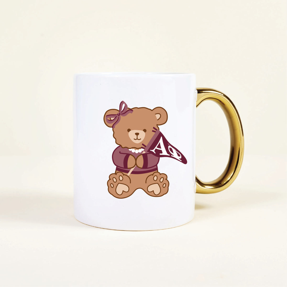 The Darling Effect’s Alpha Phi 11 oz Cozy Cute Ceramic Mug features a teddy bear with a bow and pearl necklace, holding a maroon AΦ flag—a delightful sorority mug to brighten your morning coffee.