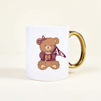 The Darling Effect’s Alpha Phi 11 oz Cozy Cute Ceramic Mug features a teddy bear with a bow and pearl necklace, holding a maroon AΦ flag—a delightful sorority mug to brighten your morning coffee.