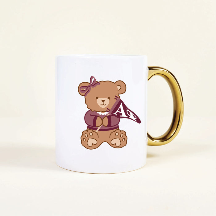The Darling Effect’s Alpha Phi 11 oz Cozy Cute Ceramic Mug features a teddy bear with a bow and pearl necklace, holding a maroon AΦ flag—a delightful sorority mug to brighten your morning coffee.