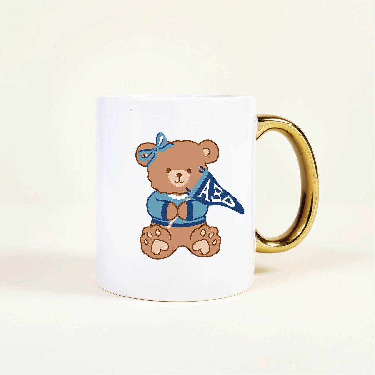 The Darling Effect’s Alpha Xi Delta 11 oz Cozy Cute Ceramic Mug features a gold handle and a brown teddy bear in a blue sweater and bow, holding an ΑΞΔ pennant—perfect for any sorority member!.