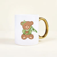 The Darling Effect’s Kappa Delta 11 oz Cozy Cute Ceramic Mug features a white base, gold handle, and an adorable teddy bear in a green KA headband, scarf, and pearls—a perfect addition to your collection.