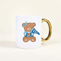 The Darling Effect’s Kappa Kappa Gamma 11 oz Cozy Cute Ceramic Mug features a white base, gold handle, and a brown teddy bear in blue clothes with Greek letters ΚΚΓ—perfect for any Kappa Kappa Gamma member.