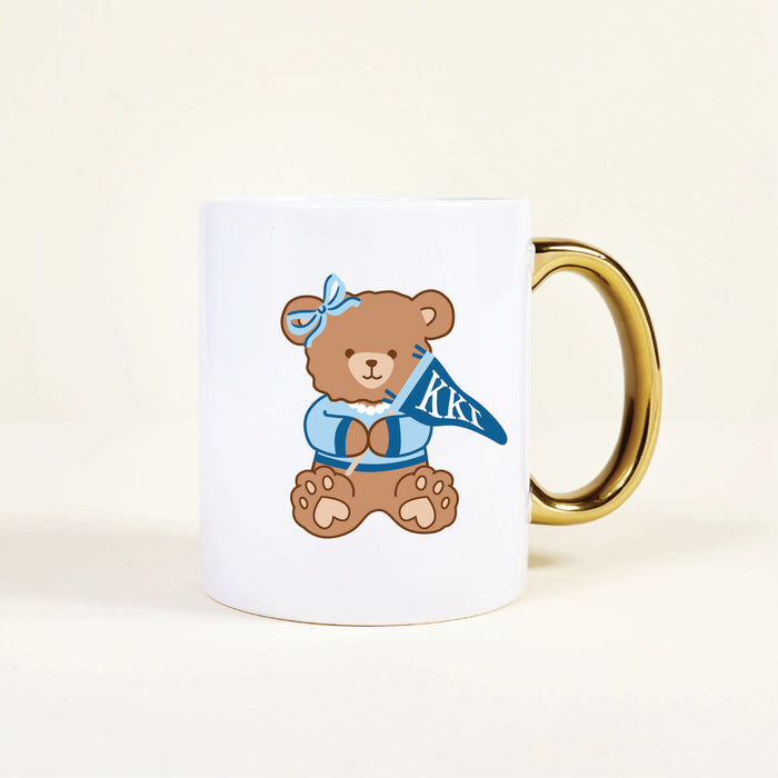 The Darling Effect’s Kappa Kappa Gamma 11 oz Cozy Cute Ceramic Mug features a white base, gold handle, and a brown teddy bear in blue clothes with Greek letters ΚΚΓ—perfect for any Kappa Kappa Gamma member.
