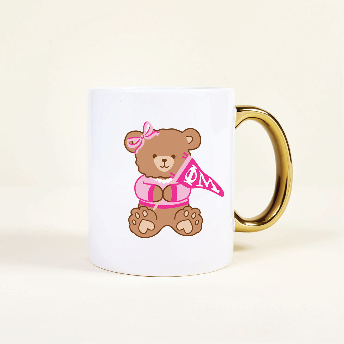 The Darling Effect’s Phi Mu 11 oz Cozy Cute Ceramic Mug is a white ceramic mug with a gold handle, featuring a brown bear in a pink bow and cardigan holding a flag with ΦM—perfect for showing sorority spirit.