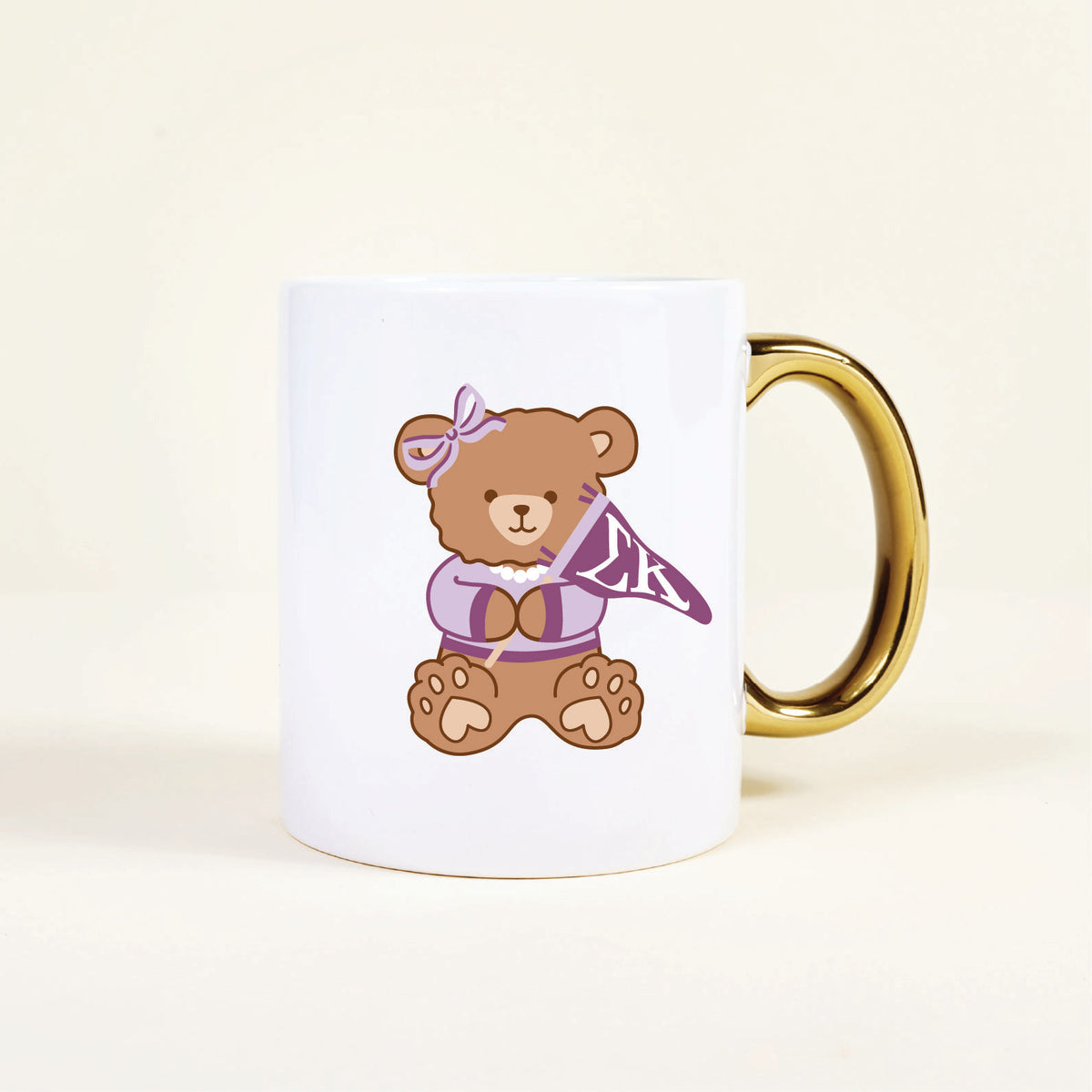 The Sigma Kappa 11 oz Cozy Cute Ceramic Mug by The Darling Effect features a brown teddy bear in a purple bow and sweater, holding an “LK” pennant. The white mug has a gold handle for stylish flair.
